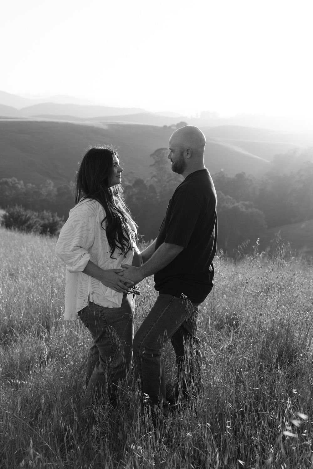 Black and white photo of a couple embracing in a grassy field with misty hills in the background.