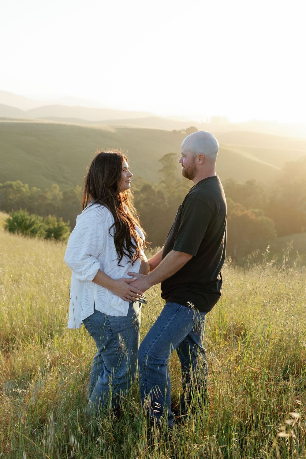 Happy pregnant couple holding hands face-to-face in tall grass field with beautiful sunlit rolling hills behind them.