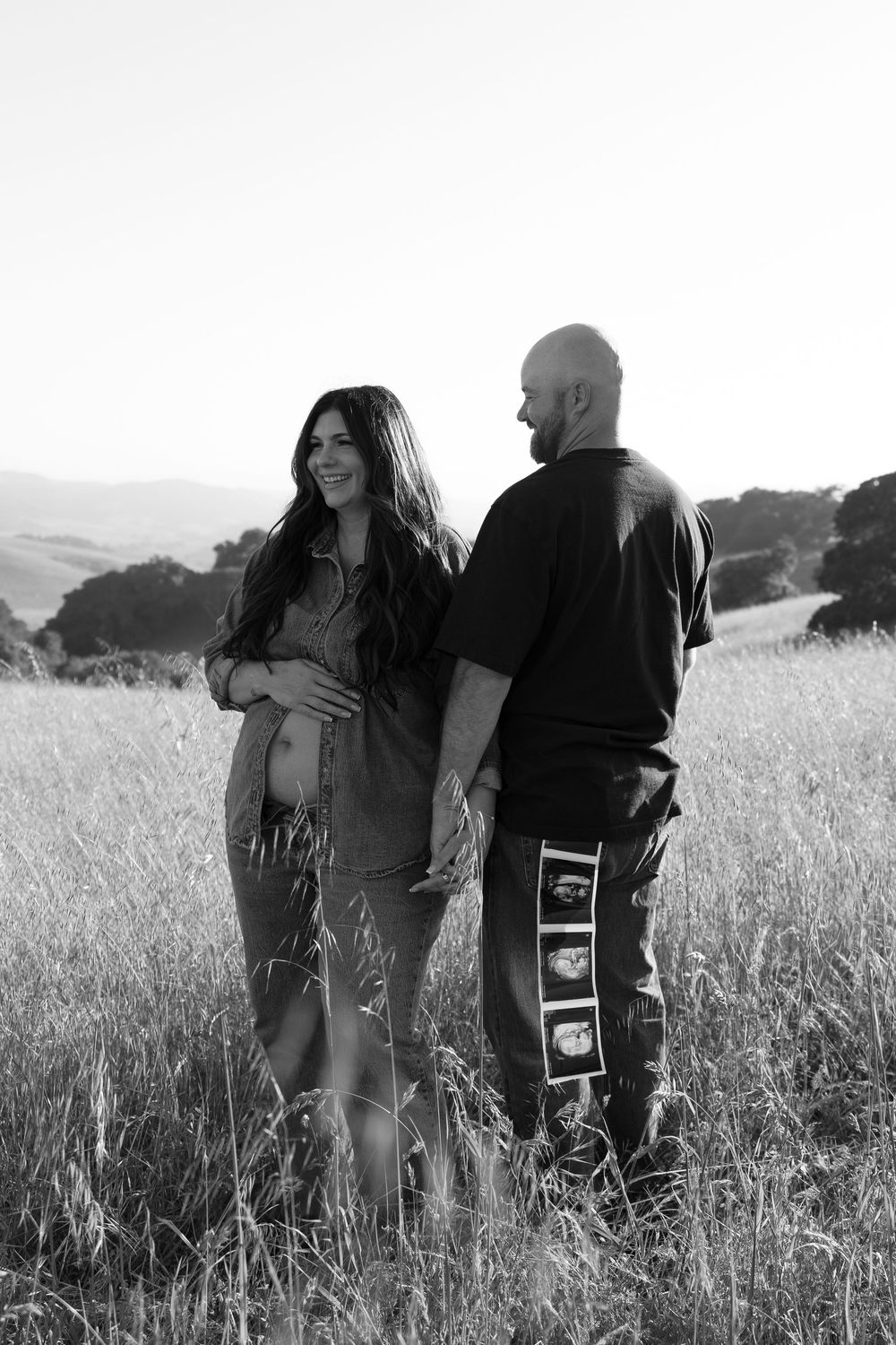 Black and white photo of pregnant couple holding hands walking through golden grass hillside field.