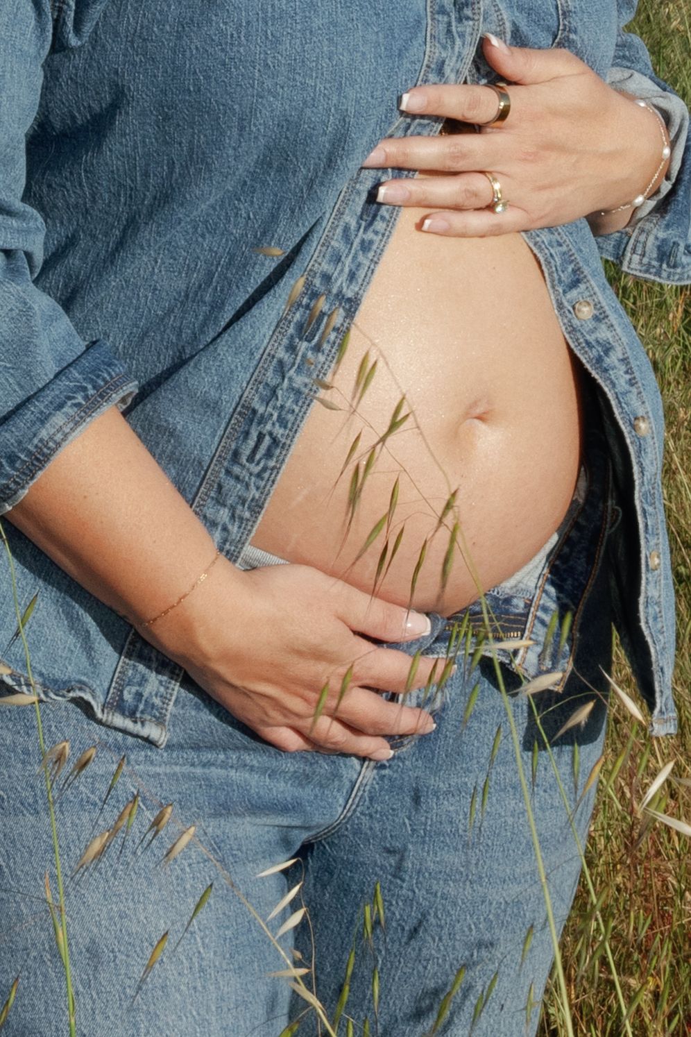 Close-up of pregnant woman in denim jacket cradling bare baby bump with grass blades nearby.