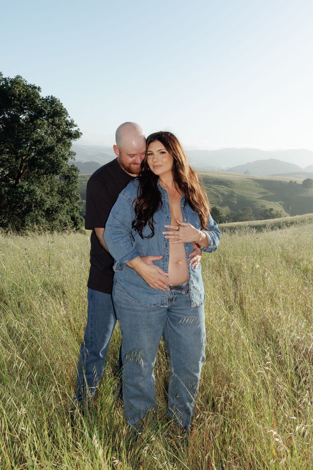 Couple poses in golden grass field, woman in denim outfit cradling baby bump, man embracing her, rolling hills backdrop.