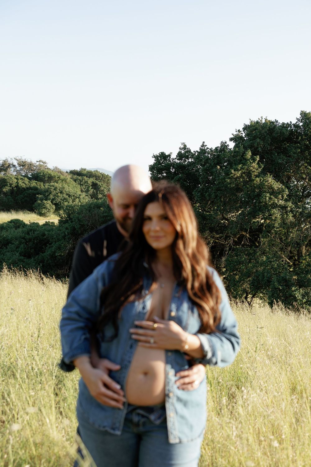 Soft-focus couple in tall grass field, man embracing pregnant woman in denim jacket showing baby bump.