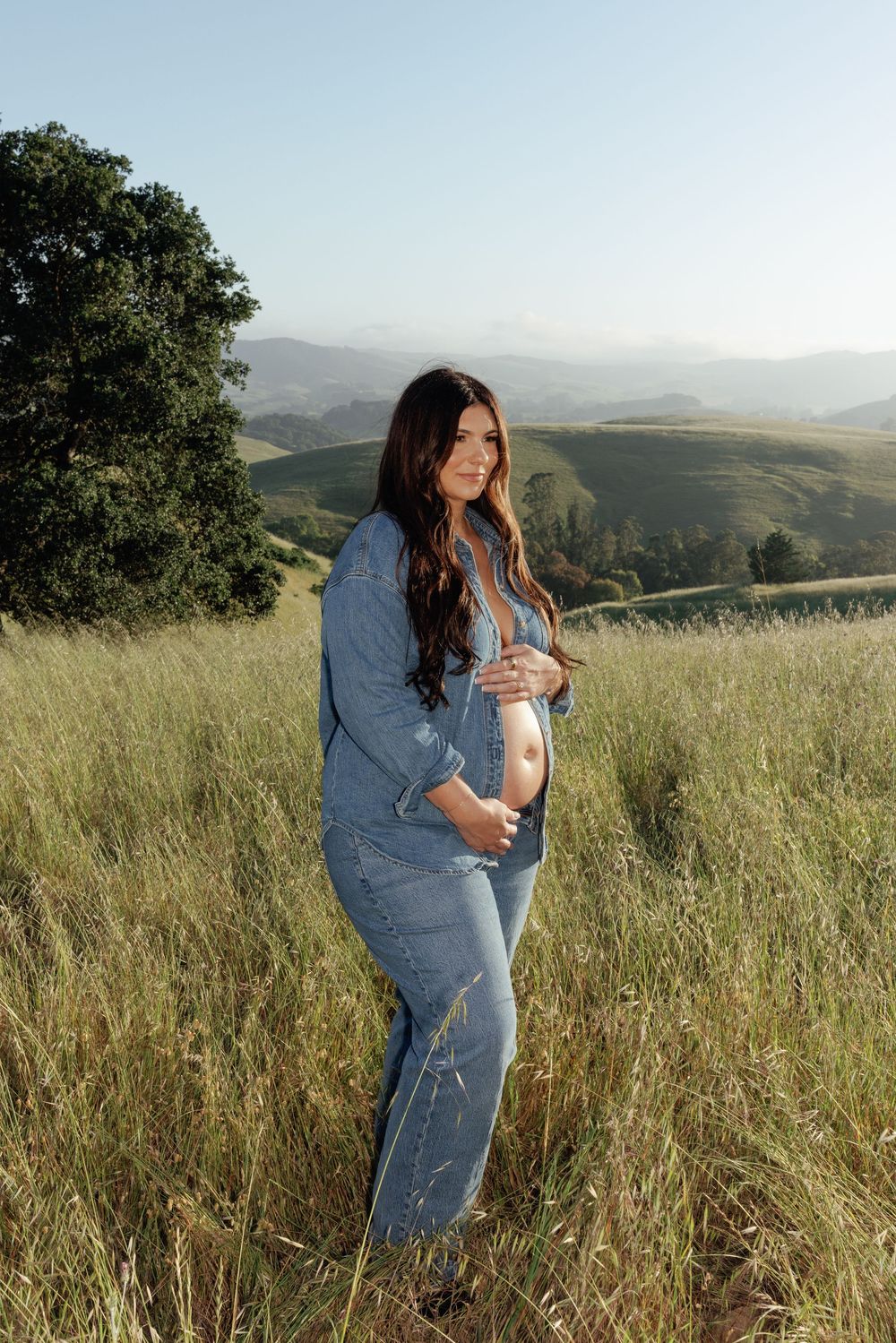 Pregnant woman in denim outfit cradling baby bump in sunlit grassy hillside field.