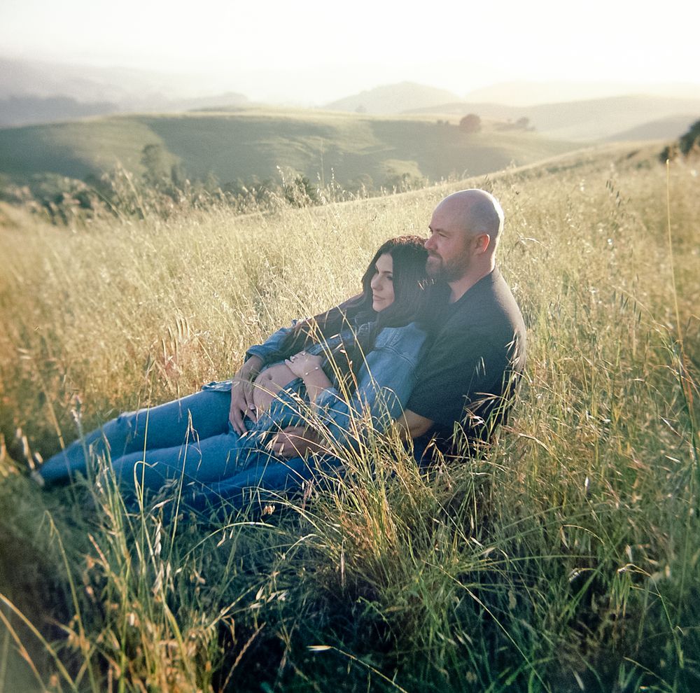 Couple sitting together in tall golden grass, embracing with scenic rolling hills in background.