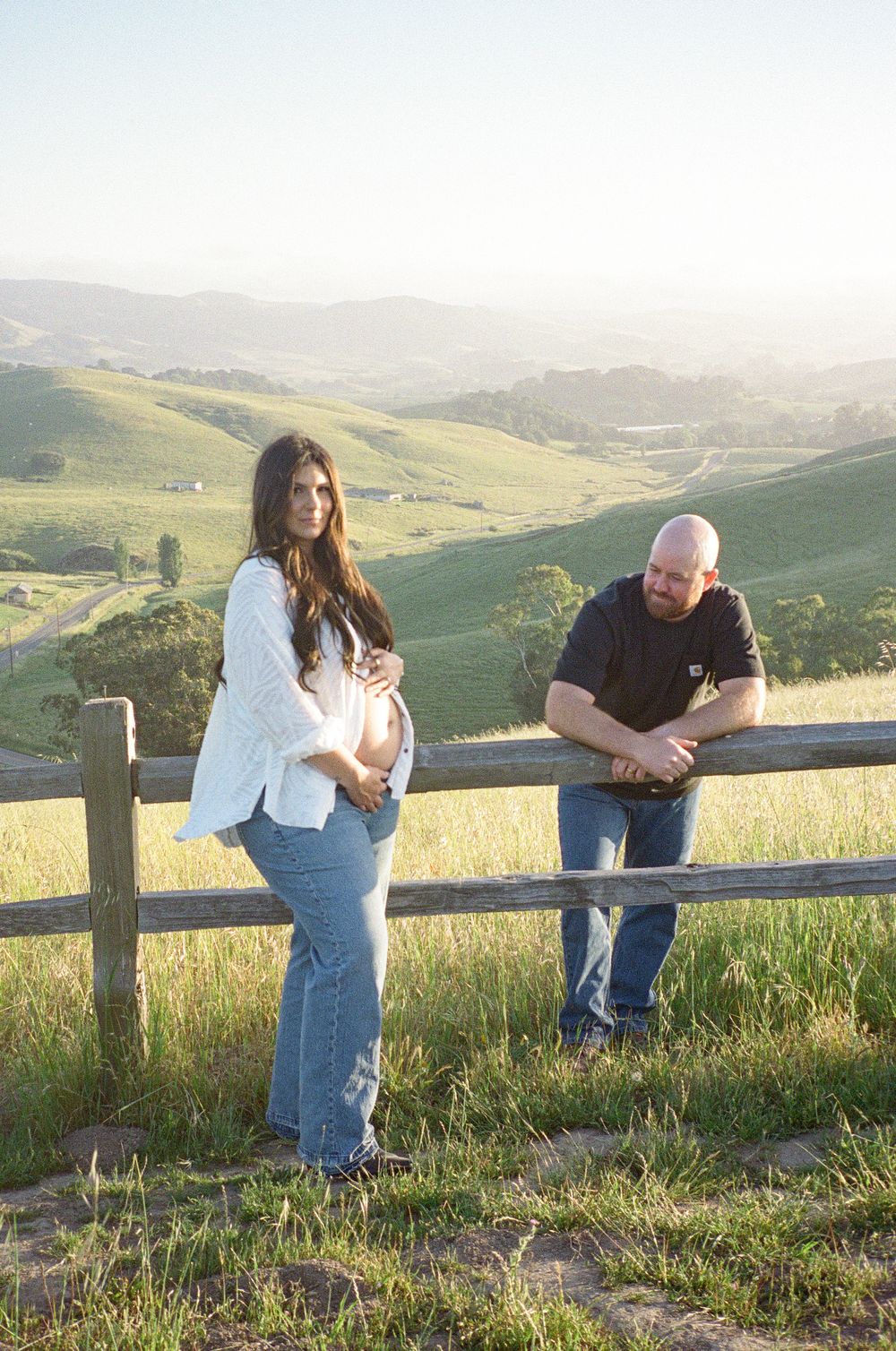 Pregnant woman leaning on rustic fence with partner beside her overlooking stunning green rolling hills at golden hour.
