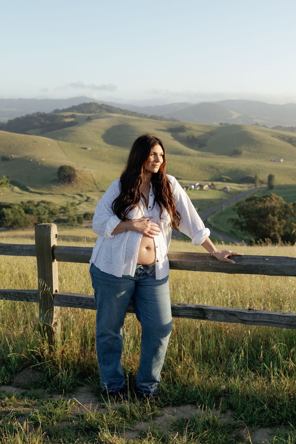 Smiling pregnant woman in open white shirt and jeans leaning on wooden fence, scenic green hillside landscape in background.
