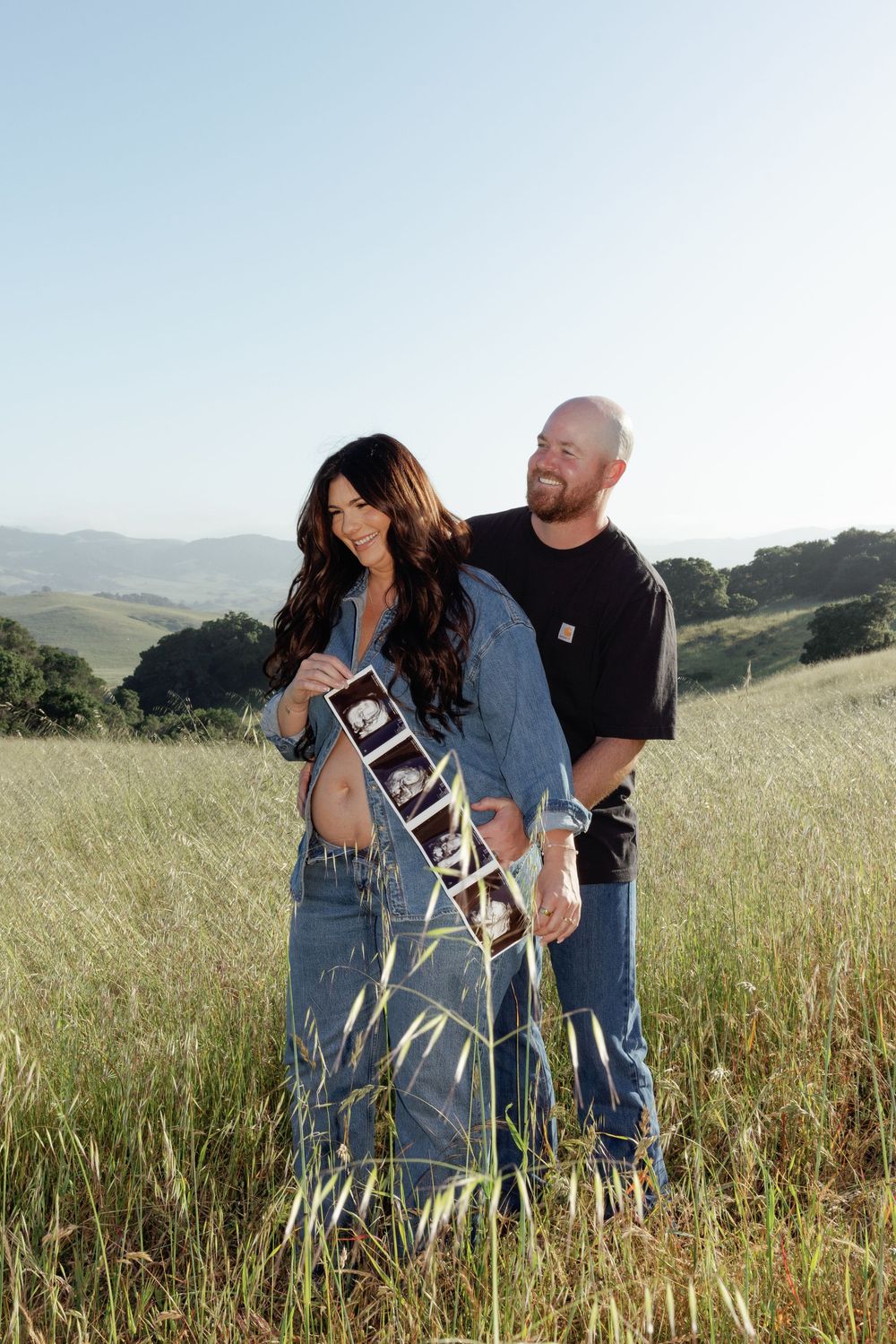 Couple standing in golden field holding ultrasound photos, pregnancy announcement with rolling hills backdrop.