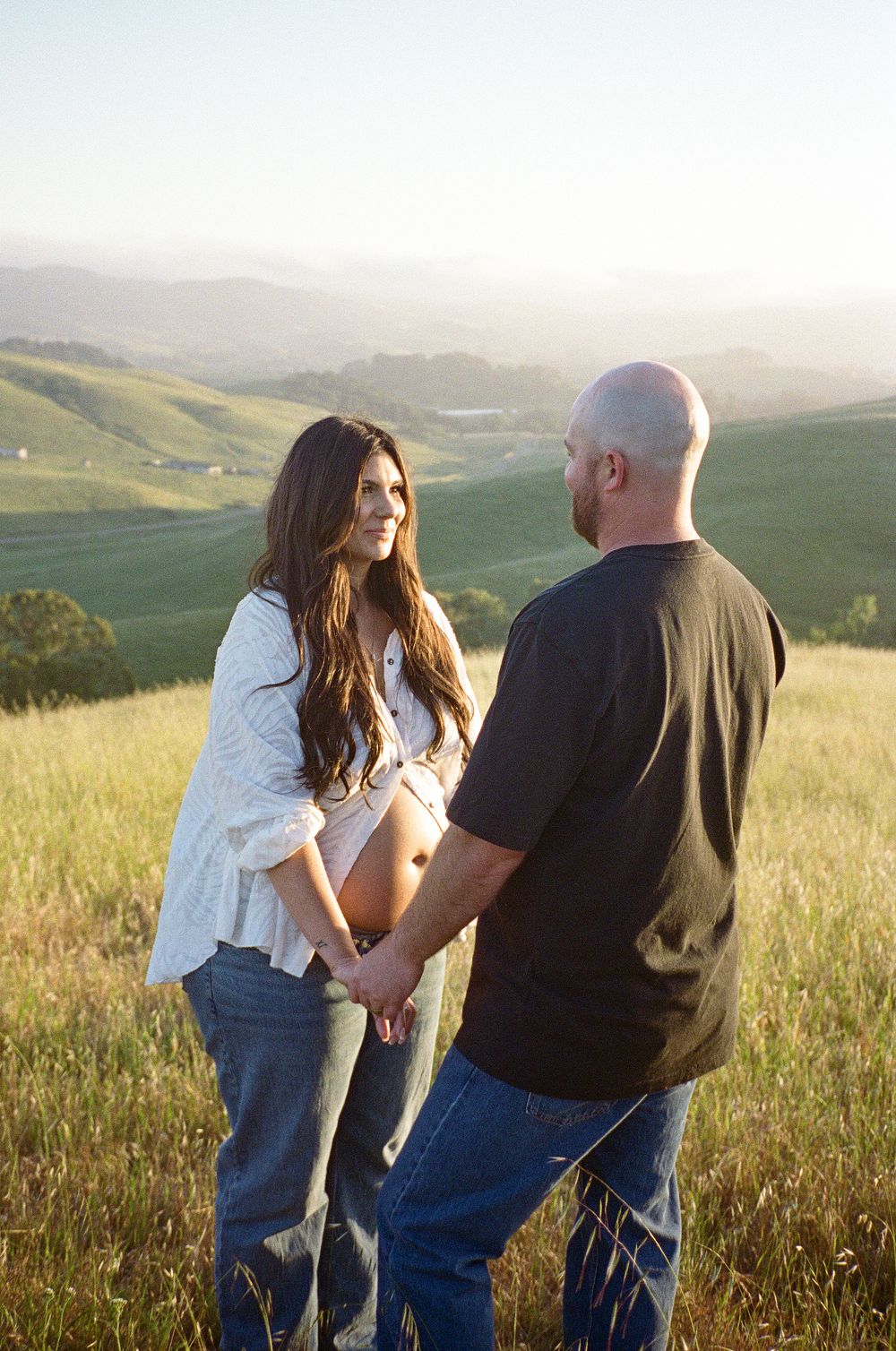 Expecting couple holds hands on a scenic hillside at golden hour, woman smiling at partner.
