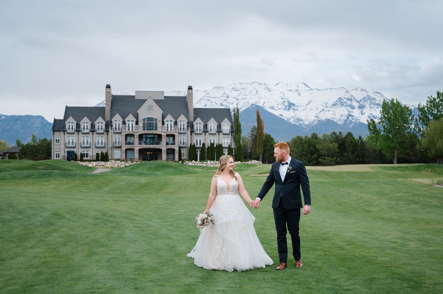 Bride and groom holding hands on lush golf course with snow-capped mountains and elegant mansion backdrop.