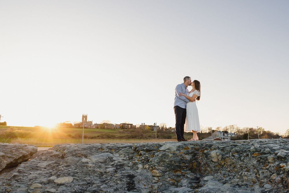 Couple embracing at sunset on rocky shore, golden light glowing behind them against a clear sky.
