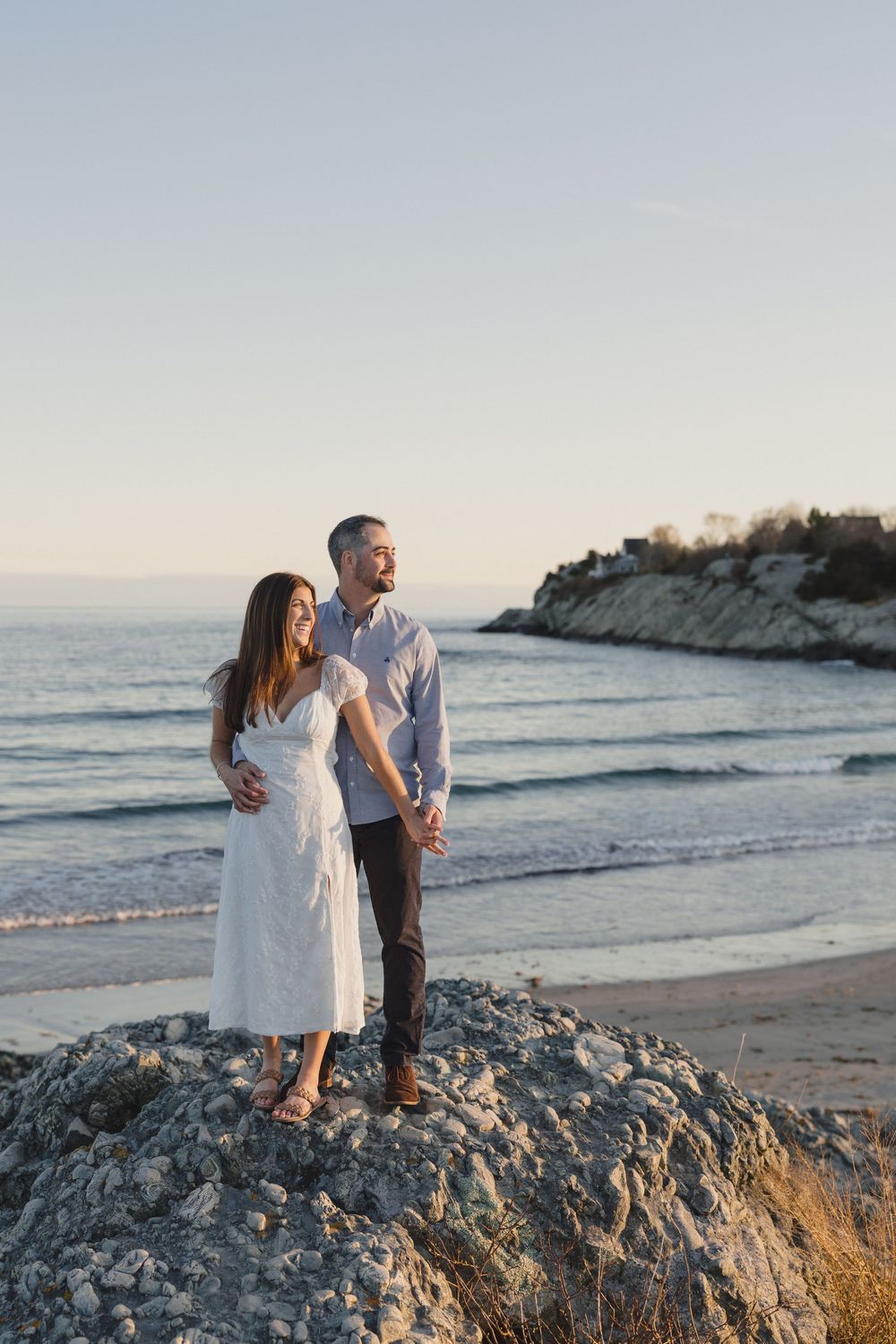 Couple standing on rocky beach at sunset, woman in white dress embraced by man in light blue shirt with ocean backdrop.