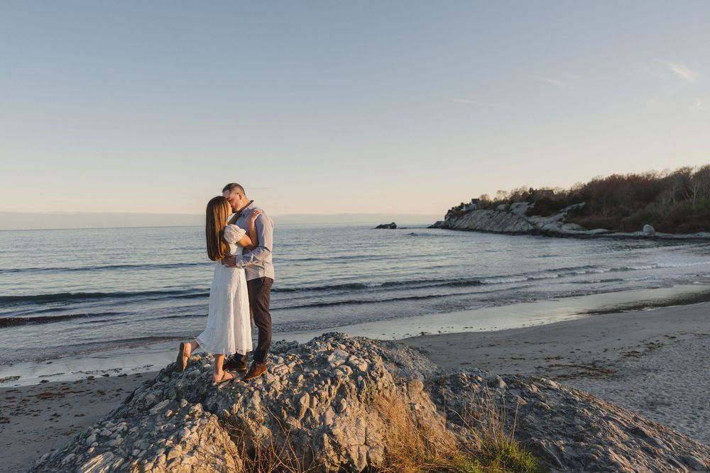 Couple sharing a romantic kiss on rocky beach at golden hour with calm ocean and coastal cliffs in background.