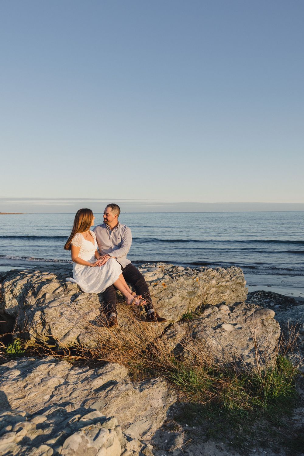 Romantic couple sitting together on coastal rocks at golden hour, gazing at each other with calm ocean behind them.