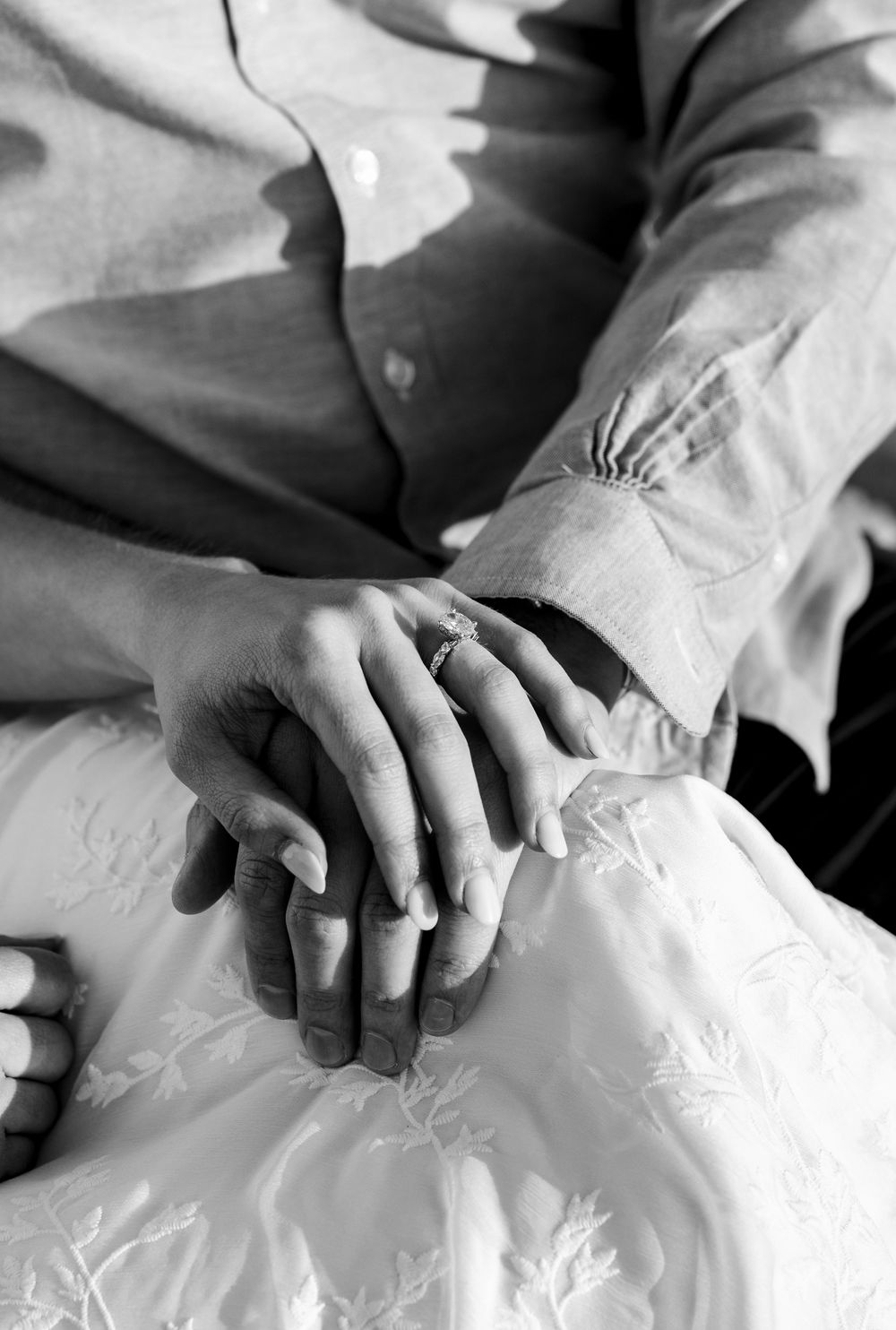 Black and white photo of a couple holding hands, bride in white dress with groom in linen shirt, wedding ring visible.
