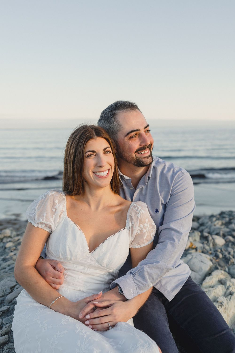 Smiling couple sitting on beach rocks at sunset, woman in white lace dress embraced by bearded man in blue shirt.
