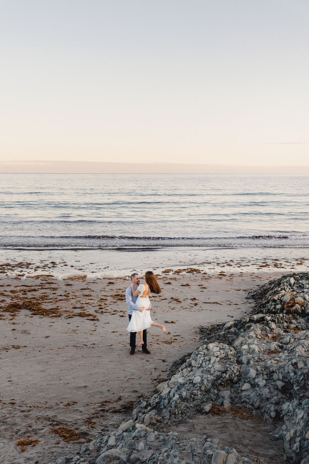 Couple embracing romantically on a rocky beach at sunset, woman in white dress, soft pastel sky and ocean backdrop.