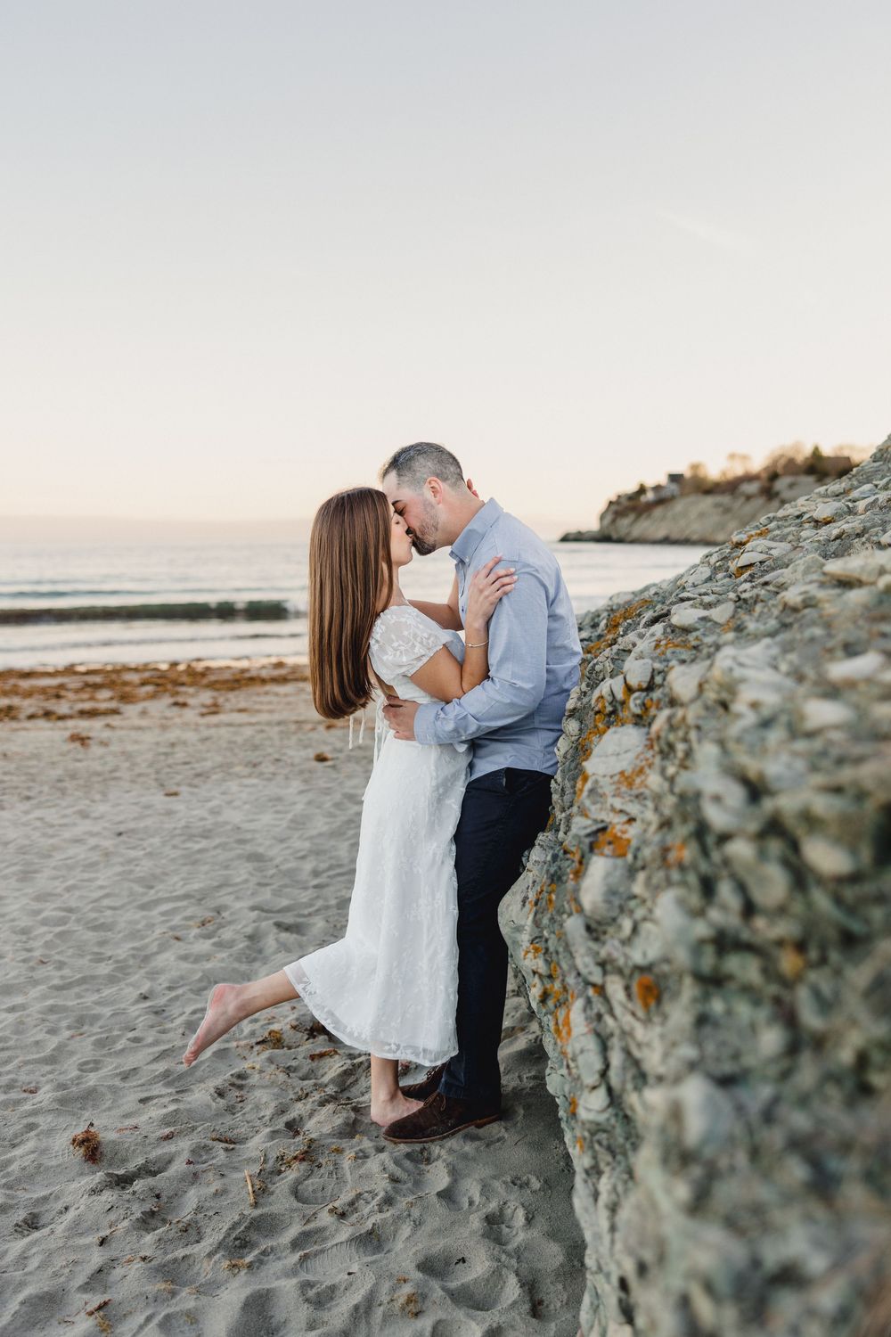 Romantic couple kissing against a rocky cliff on a sandy beach at sunset.