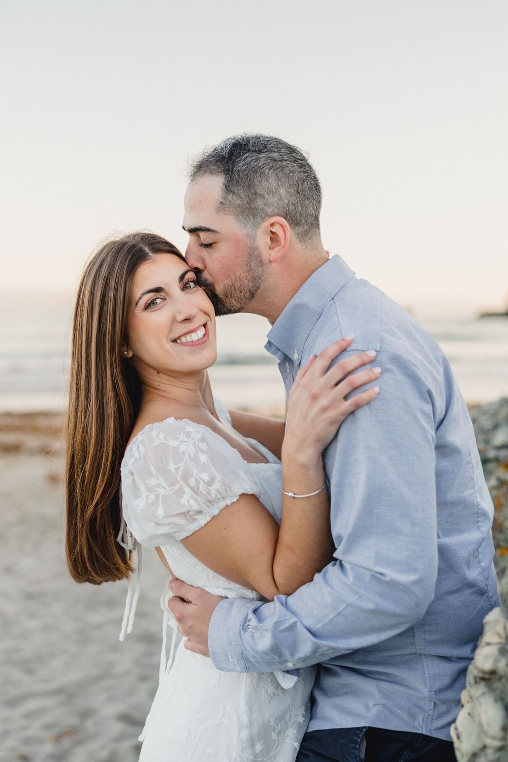 Man kissing woman on cheek at beach during golden hour, couple embracing romantically by the ocean.