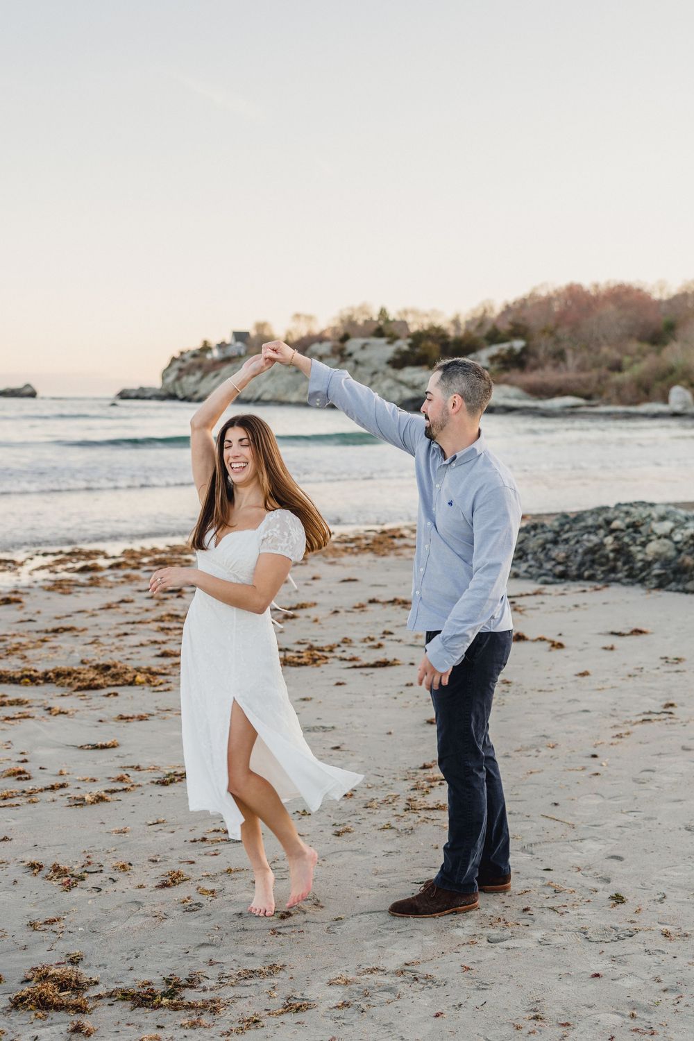Couple dancing on a sandy beach at sunset, woman in white dress being twirled by man in blue shirt.