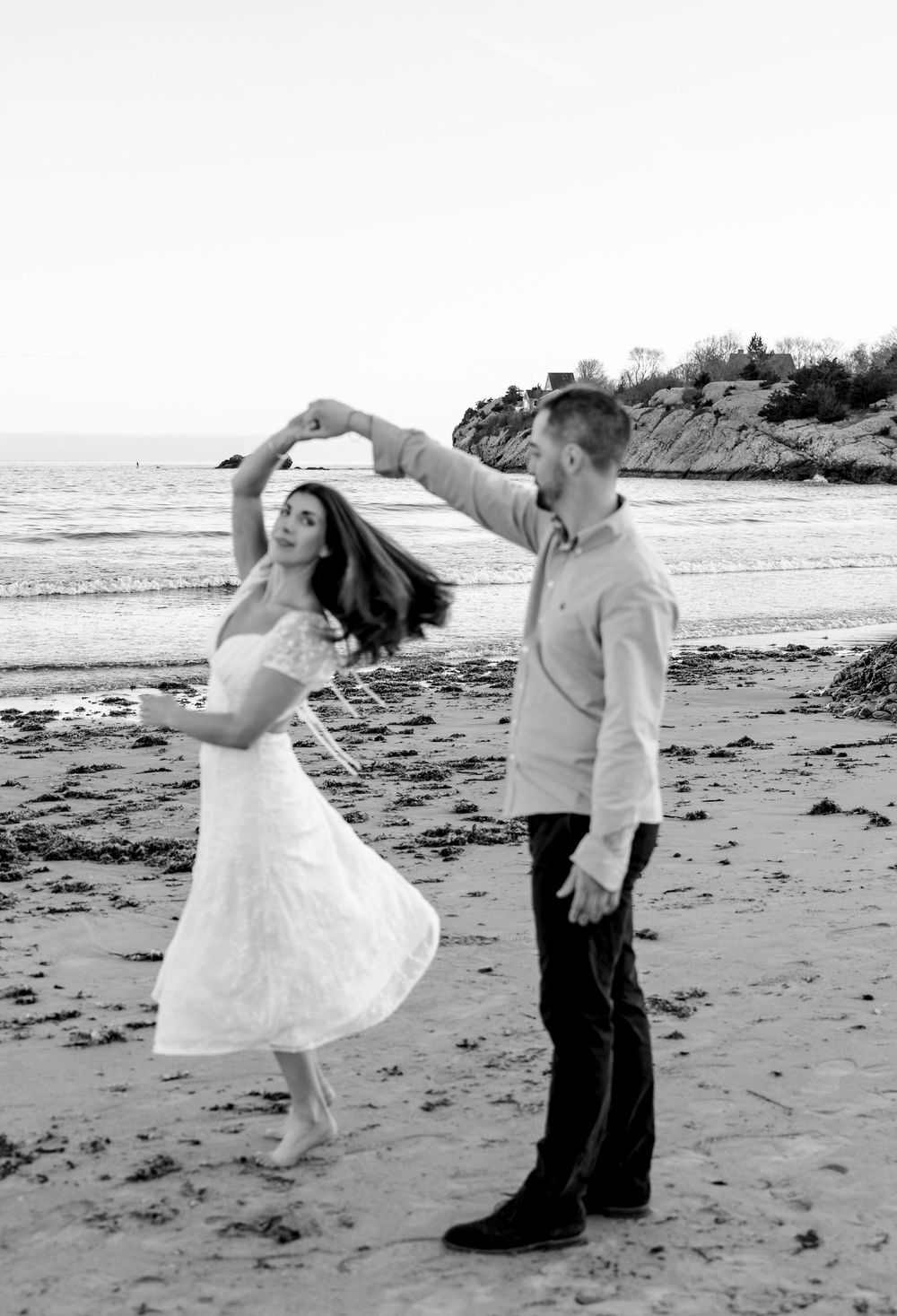 Couple dancing on beach, man spinning woman in white dress, black and white photo, ocean backdrop.