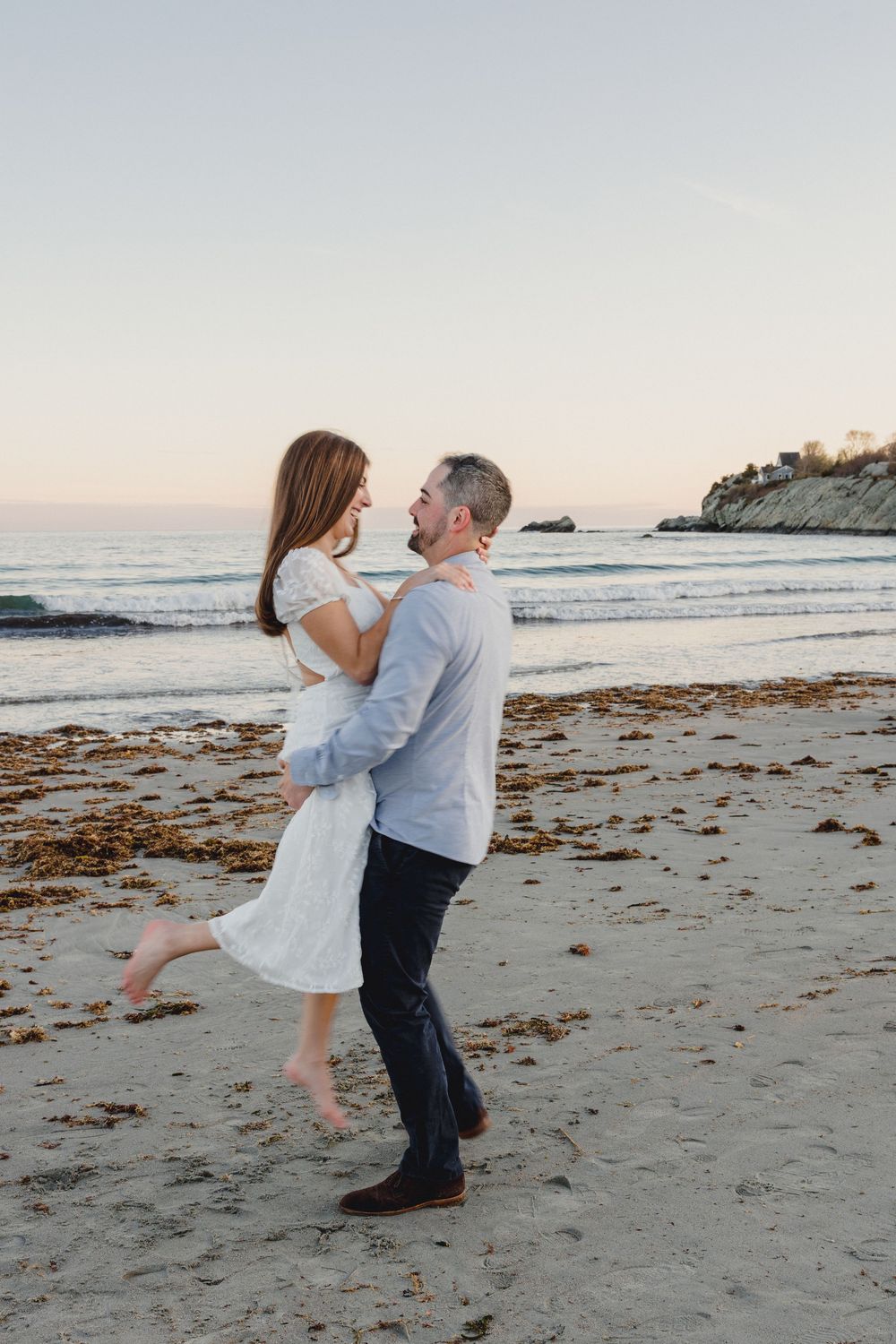 Romantic couple embracing on a beach at sunset, woman in white dress lifted by man in blue shirt.
