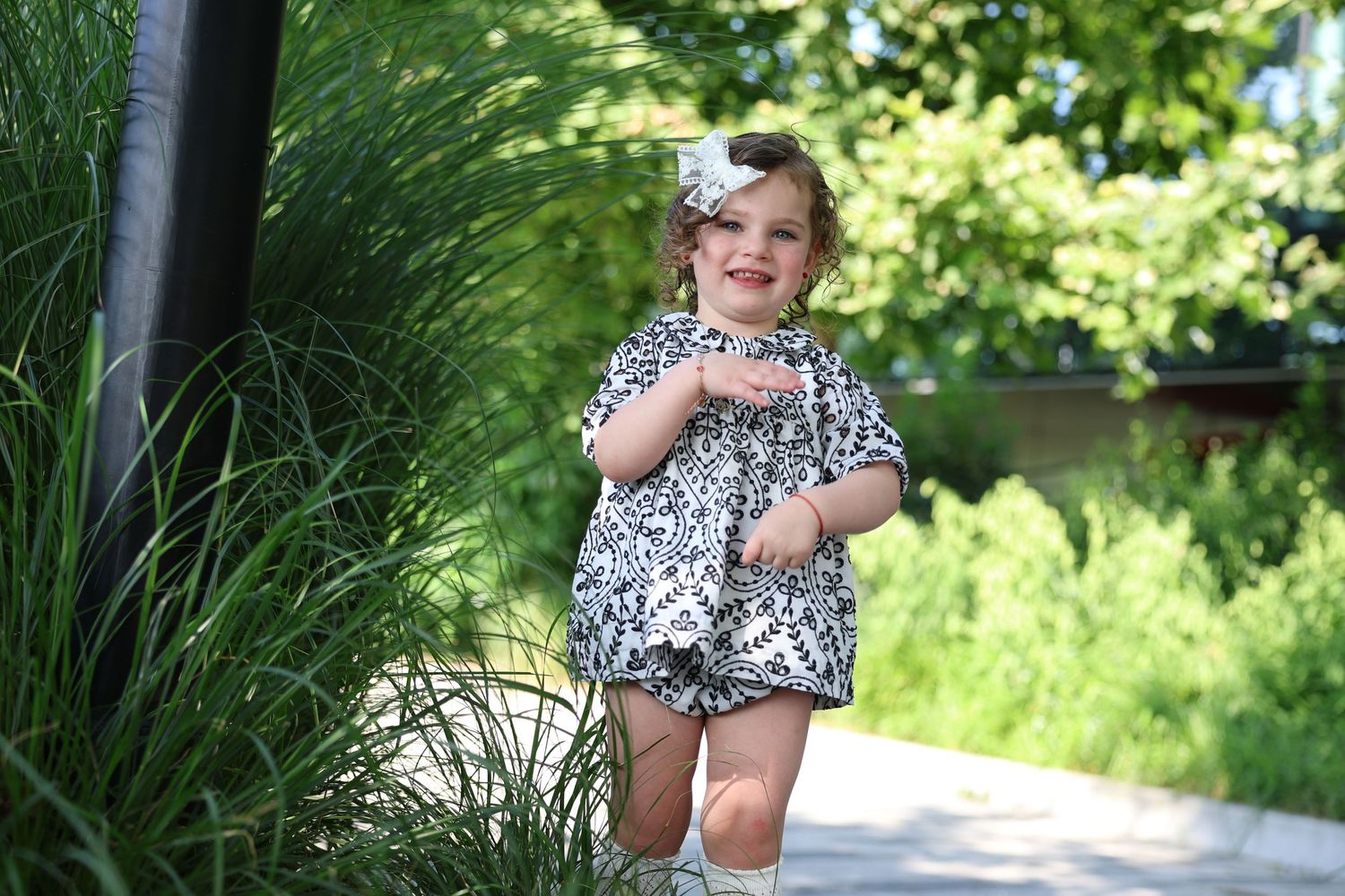 A happy toddler girl in a floral outfit walks along a garden path surrounded by tall green plants.
