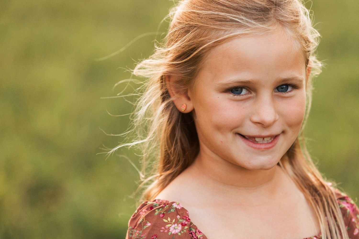 A young girl in a burgundy dress smiles while standing outdoors in natural sunlight.