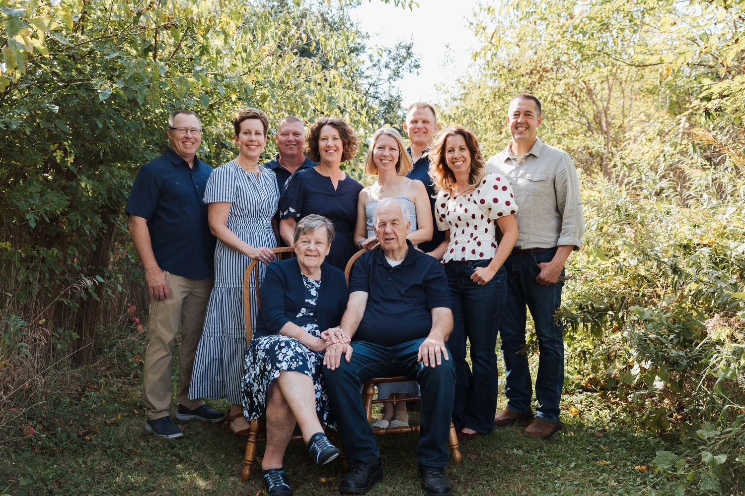 A multi-generational family group poses together outdoors on a sunny day with green foliage in the background.
