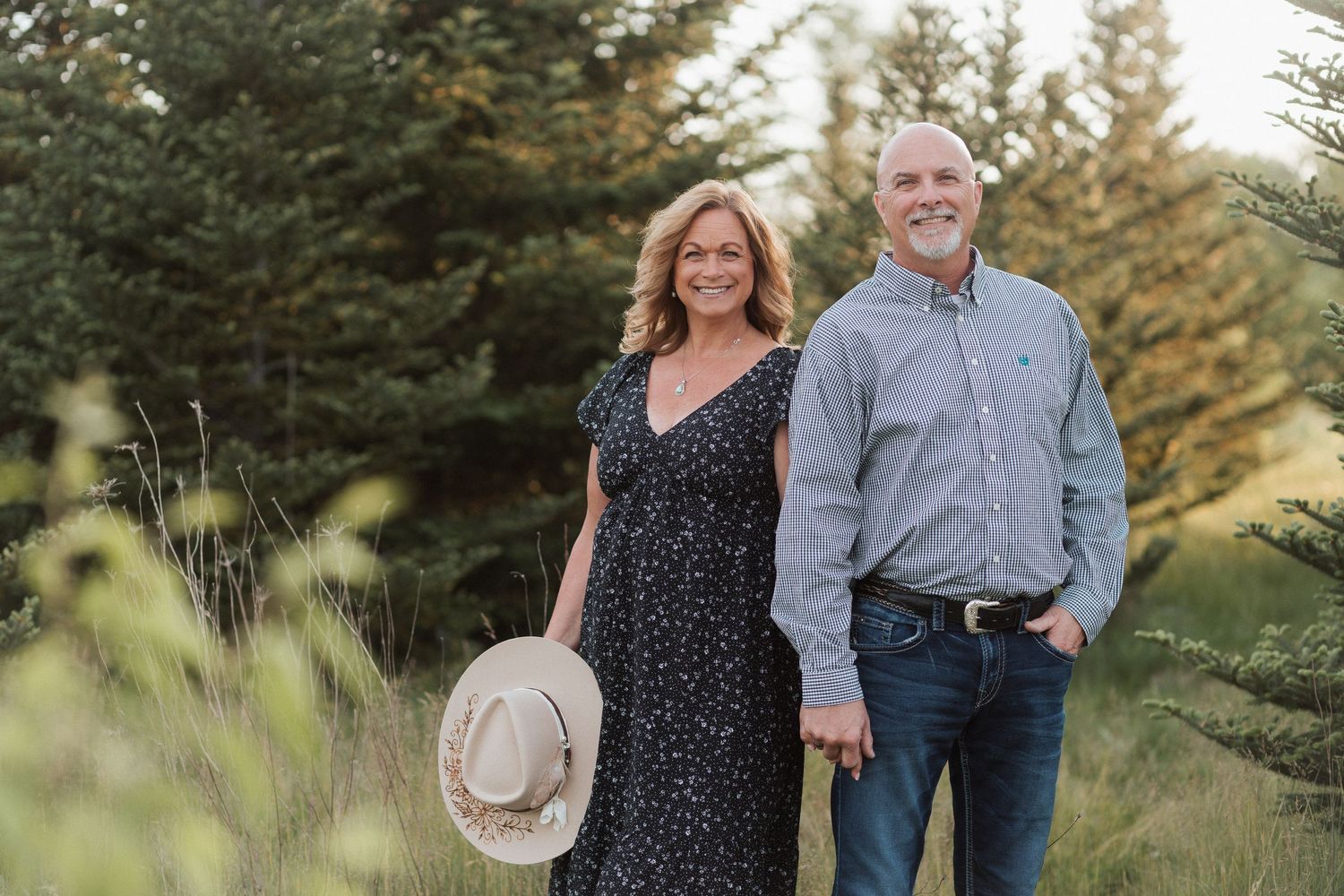 A smiling couple holds hands outdoors near evergreen trees, woman in floral dress holding hat, man in blue shirt.