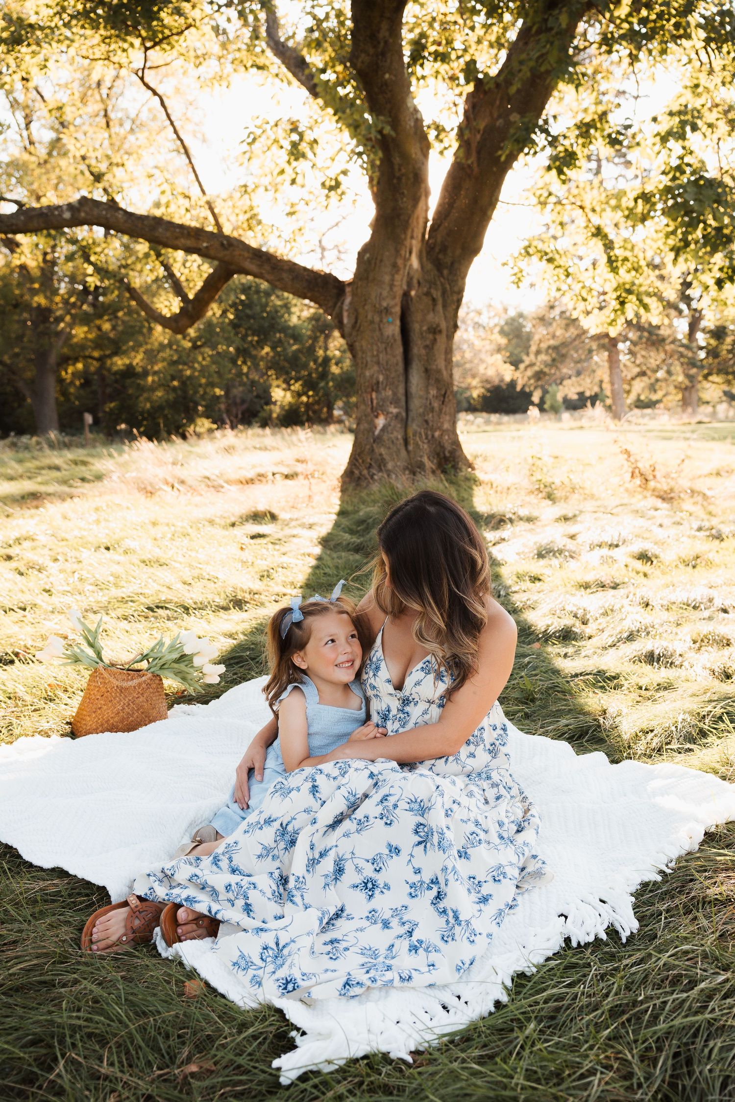Two people in matching blue floral dresses sit together on a white blanket under a sunlit tree in a grassy field.