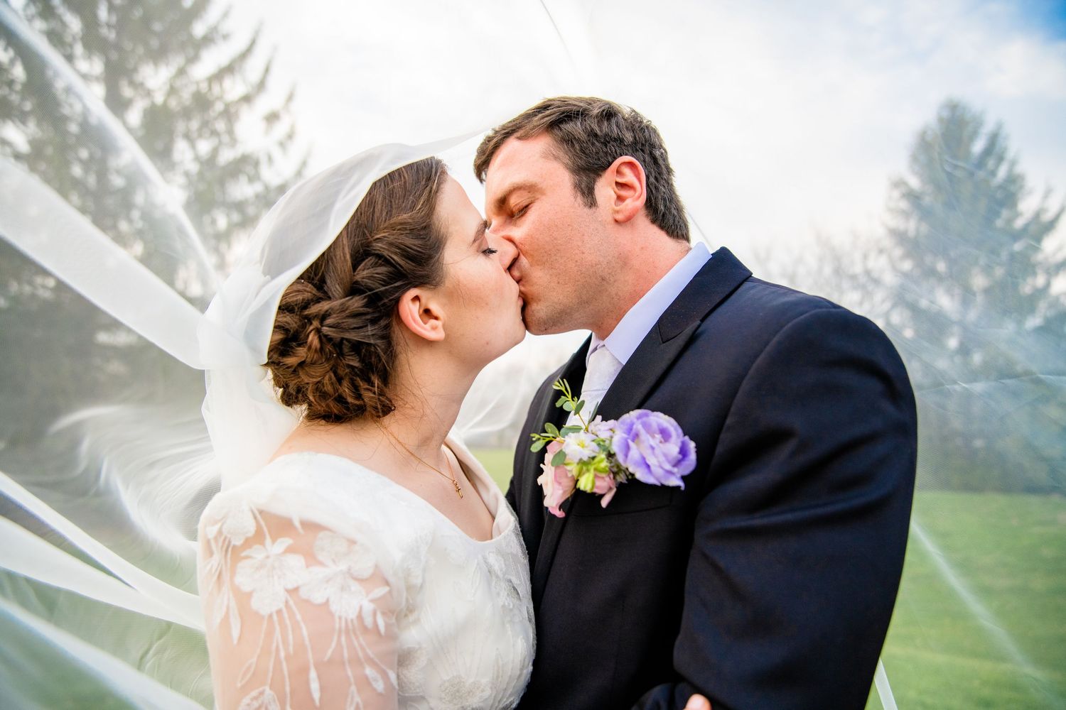 Bride and groom sharing a romantic kiss under a flowing white veil outdoors on their wedding day.