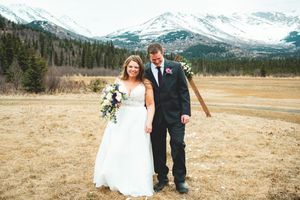 Bride and groom smiling together in a mountain meadow, bride holding a bouquet, snow-capped peaks in the background.