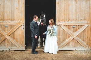 Bride and groom stand with a black horse in front of rustic wooden barn doors on their wedding day.
