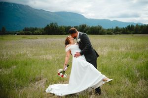 Bride and groom share a romantic dip kiss in a lush green meadow with misty mountains in the background.