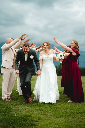 Bride and groom walk through a celebratory arch of raised hands with wedding party on a green lawn under cloudy skies.
