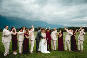 Wedding party celebrates outdoors with mountains and dramatic sky, bridesmaids in burgundy dresses and groomsmen in tan suits.