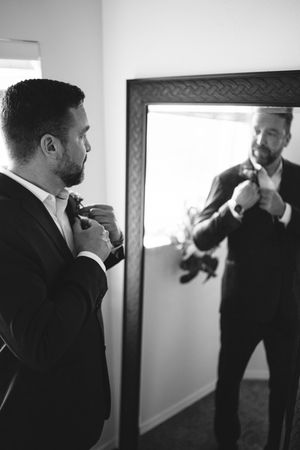 Black and white photo of a groom adjusting his boutonniere while looking in a mirror before his wedding.