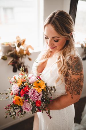 Beautiful tattooed bride in white lace dress holding a vibrant colorful wedding bouquet indoors.