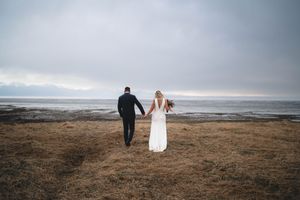Bride and groom walking hand in hand across a coastal field under a dramatic cloudy sky.