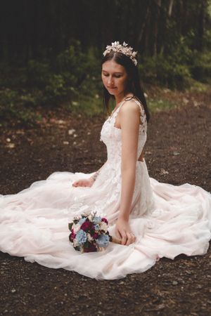 Bride in blush pink lace gown sitting on ground with bouquet, wearing floral crown in forest.
