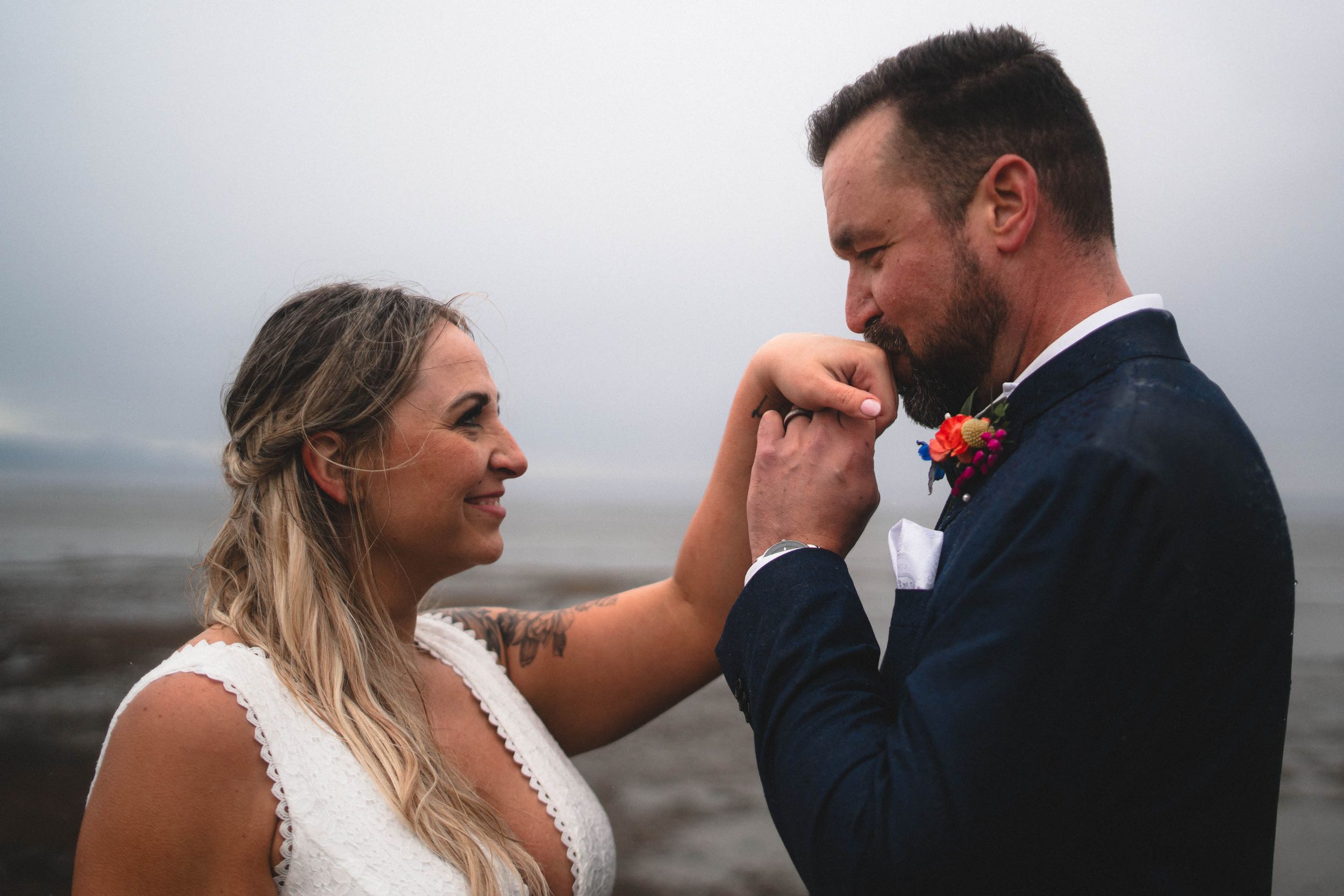 A groom tenderly kisses his brides hand on a misty beach during their intimate outdoor wedding ceremony.