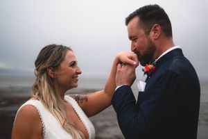 A groom tenderly kisses his brides hand on a misty beach during their intimate outdoor wedding ceremony.