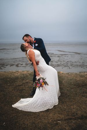 Groom dips bride for a romantic kiss by the seaside, bride in lace gown holding vibrant bouquet, misty backdrop.