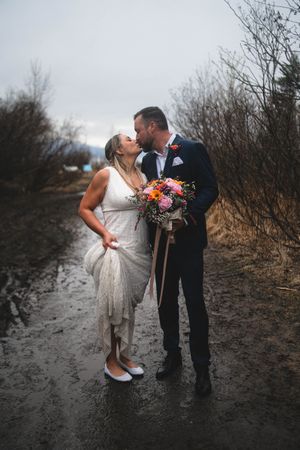 Bride and groom share a romantic kiss on a muddy path, bride holding colorful bouquet, overcast winter wedding day.