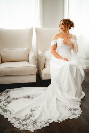 Elegant bride in off-shoulder lace wedding gown with long train sits gracefully in a white armchair by a bright window.