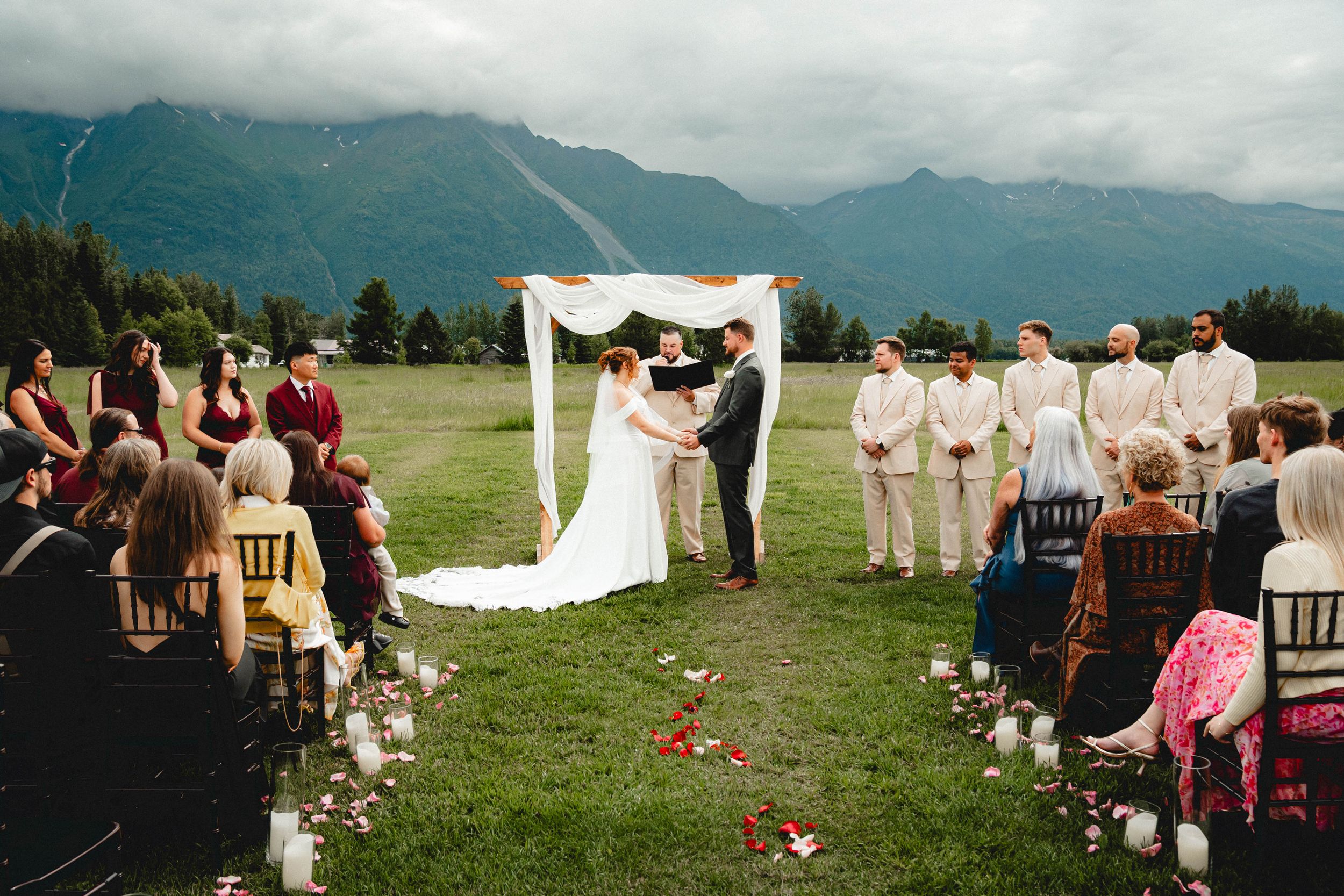 Outdoor wedding ceremony with couple at floral arch, groomsmen in tan suits, mountain backdrop, and rose petal aisle.
