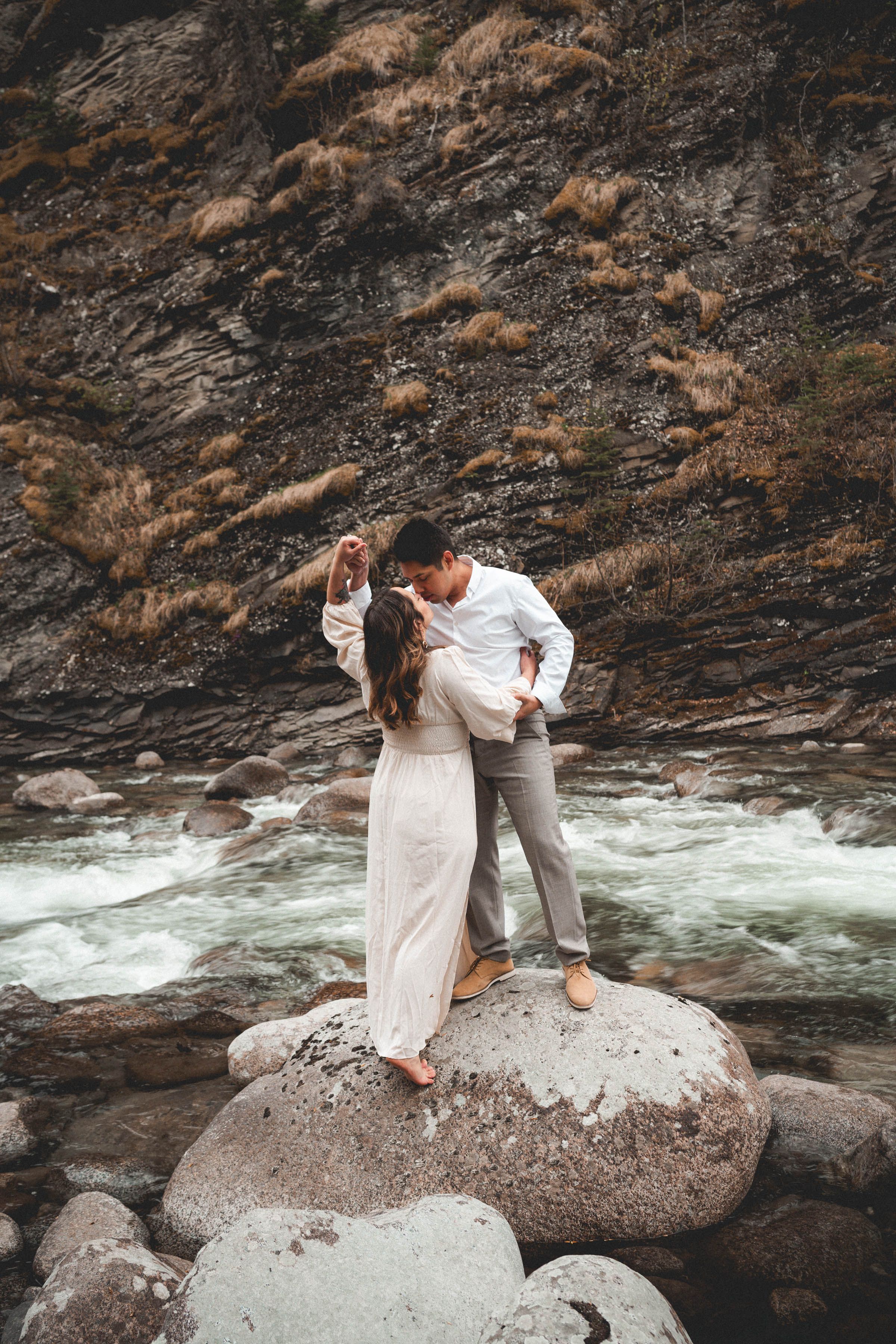 Couple dancing romantically on rocks by a rushing mountain river, bride in flowing white dress.