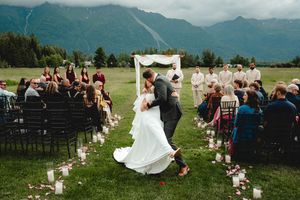 Bride and groom share first kiss at outdoor mountain wedding ceremony with guests seated along flower-lined aisle.