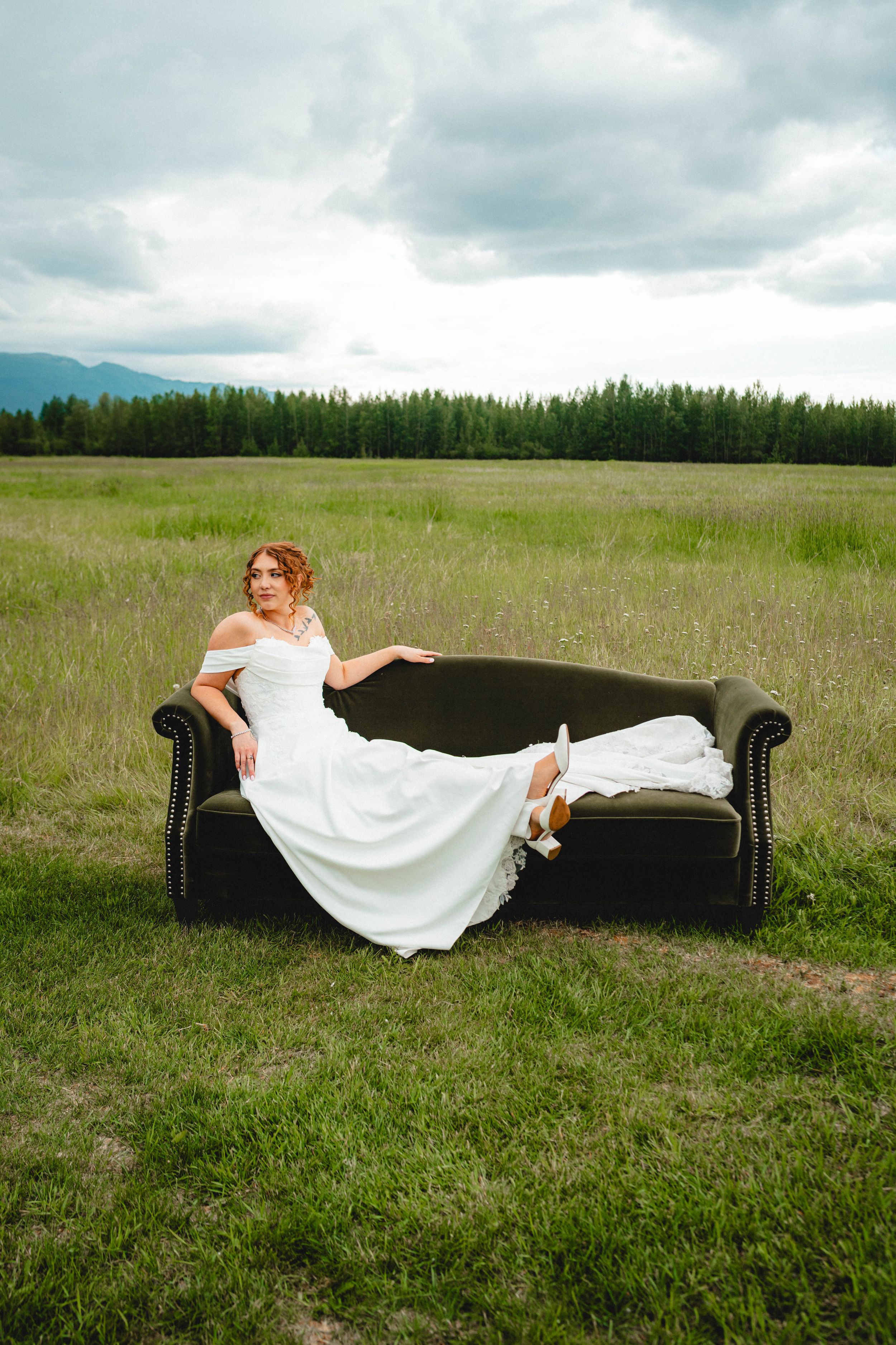 Bride in white off-shoulder gown reclines on dark leather sofa in open field with mountain forest backdrop.