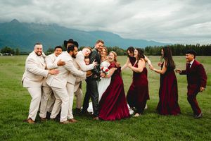 Wedding party celebrates outdoors with mountains backdrop, bridesmaids in burgundy gowns and groomsmen in tan suits.